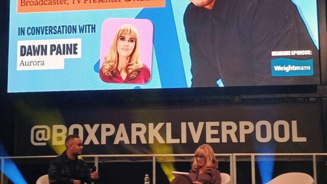 Ashley John Baptiste on stage at Raise Up, sat down in conversation with a female presenter. The screen reads 'fireside chat'