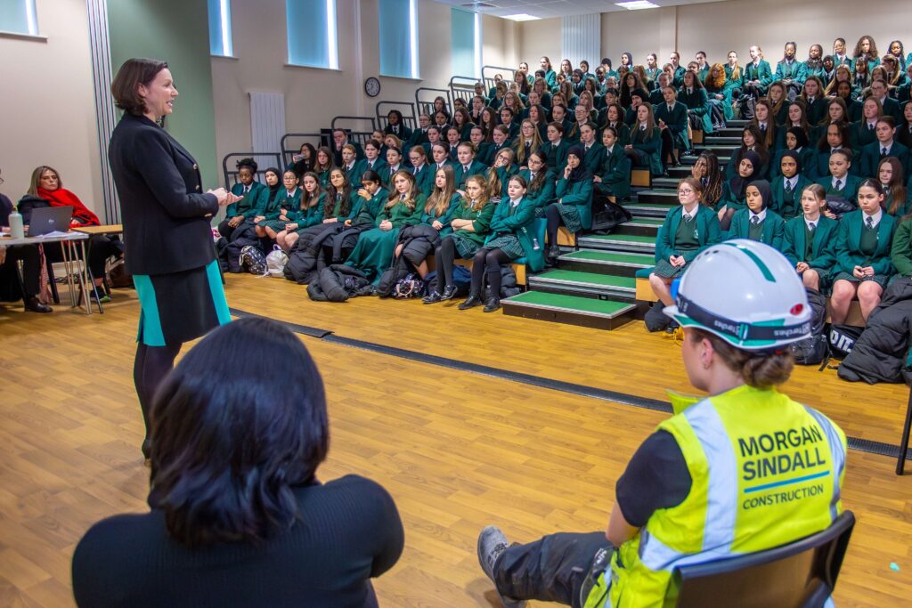A woman standing in a sports hall, talking to an assembly of schoolgirls about her career, as part of their careers curriculum.
