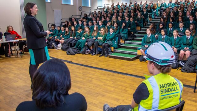 A woman standing in a sports hall, talking to an assembly of schoolgirls about her career, as part of their careers curriculum.