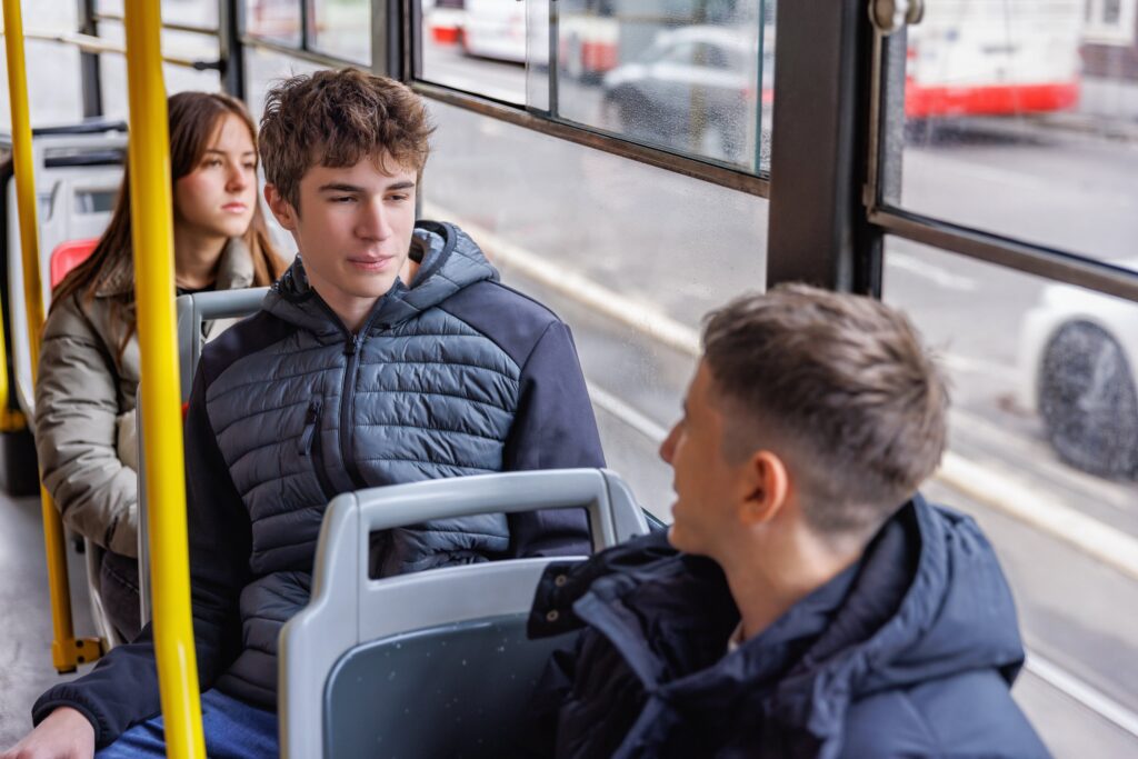 Two young men sitting on a bus. The one in front has turned to talk to the one behind. Sitting behind them both, a young woman is looking out of the window.