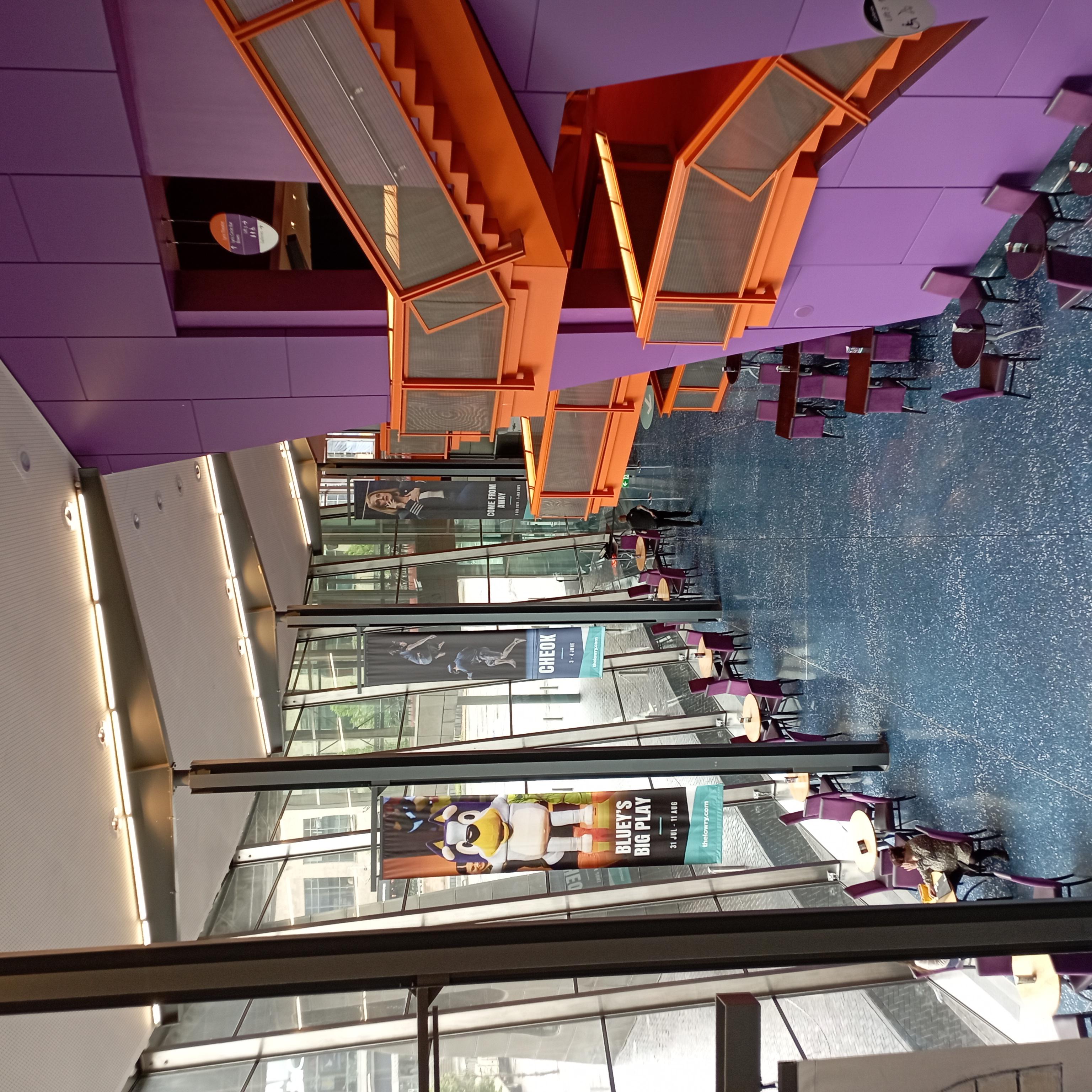 A shot from above of the Lowry main space. Purple walls, wooden banisters, and blue floors. There are banners advertising the Bluey show, Cheok dance, and Come From Away. Sunlight is streaming into the space from the large windows. 