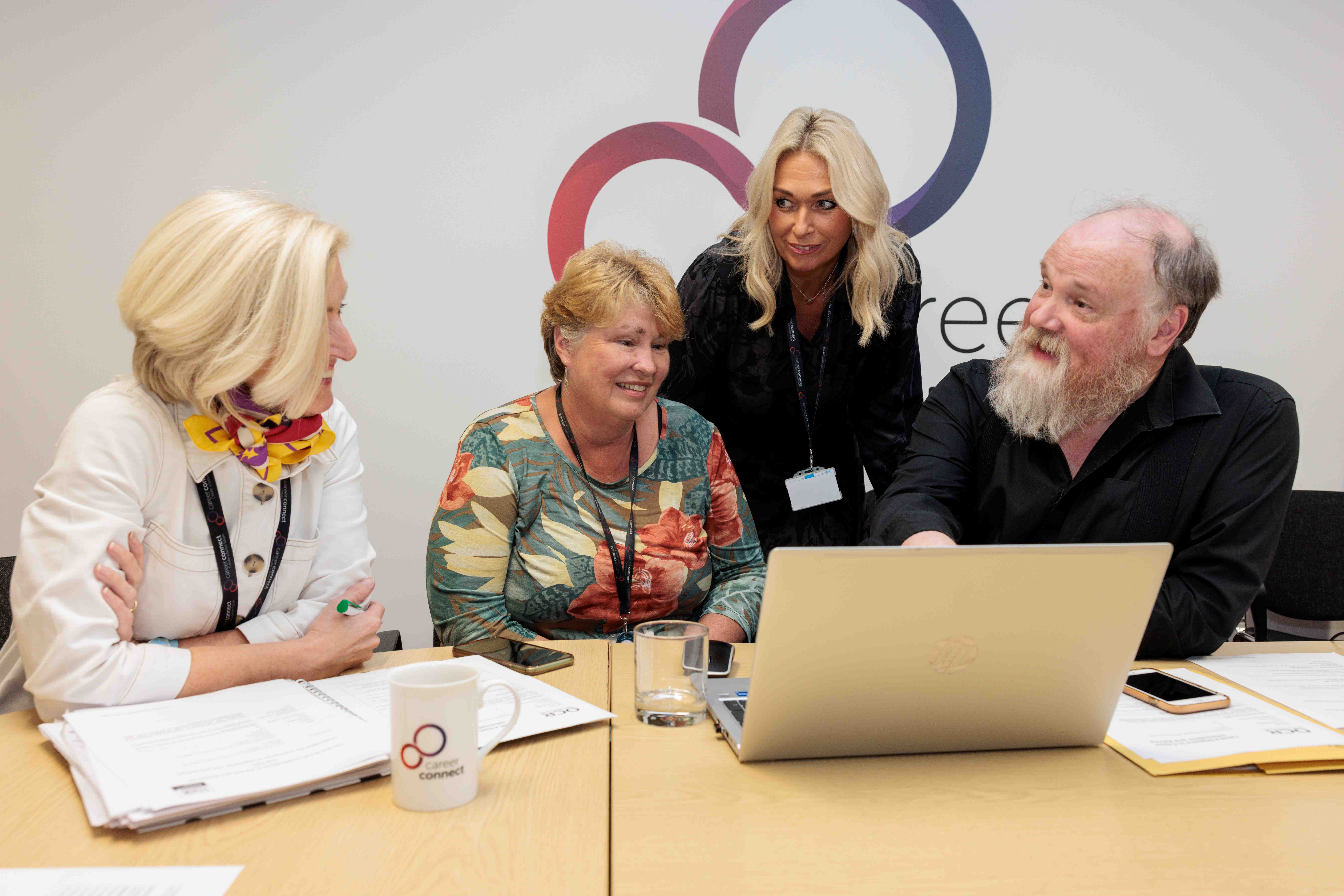 Three female and one male member of Career Connect staff sat at a desk with a laptop discussing work at an informal meeting. There is a cup on the desk.