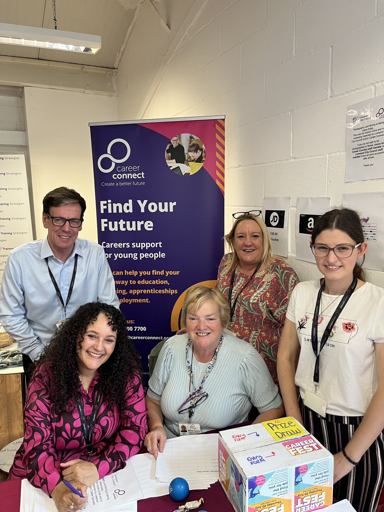 Five members of the Career Connect Liverpool team sat at a table at the entrance to the Careers Fest event. They are smiling and waiting to welcome people. 